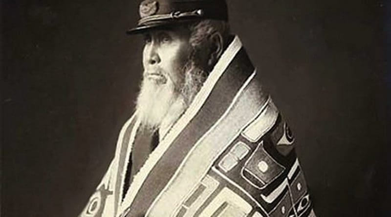 Chief Anotklosh of the Taku nation. He wears a woven Chilkan blanket of cedar bark and mountain goat wool and a European-style cape, and holds a carved wooden bird rattle. Photograph by W.H. Case, ca. 1913, Juneau, Alaska, Wikipedia Commons.