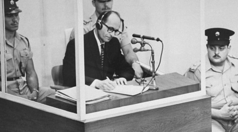Defendant Adolf Eichmann takes notes during his trial in Jerusalem. The glass booth in which Eichmann sat was erected to protect him from assassination. (Photo credit: Israeli Government)