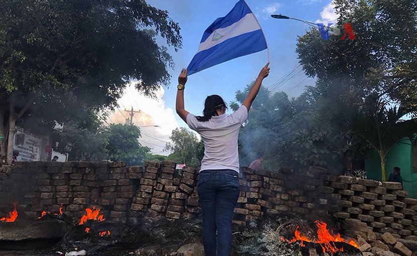A woman stands near a burning barricade holding the national flag of Nicaragua. Photo Credit: VOA