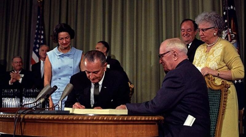 President Lyndon B. Johnson signing the Medicare Bill at the Harry S. Truman Library in Independence, Missouri. Former president Harry S. Truman is seated at the table with President Johnson. The following are in the background (from left to right): Senator Edward V. Long, an unidentified man, Lady Bird Johnson, Senator Mike Mansfield, Vice President Hubert Humphrey, and Bess Truman. Photo Credit: Lyndon Baines Johnson Library (Austin, Texas), Wikimedia Commons.