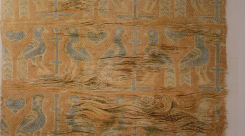 The motive is birds, probably peacocks, flanking a stylized tree or cross. It consists of several silk pieces sewn together. One piece, 30x40 cm, covers the front of the pillow and about a third of the back, while the rest consists of strips about 5 cm wide, cut off without regard to the pattern. The colors are golden and two light blue shades. CREDIT: © Nationalmuseet / The National Museum of Denmark