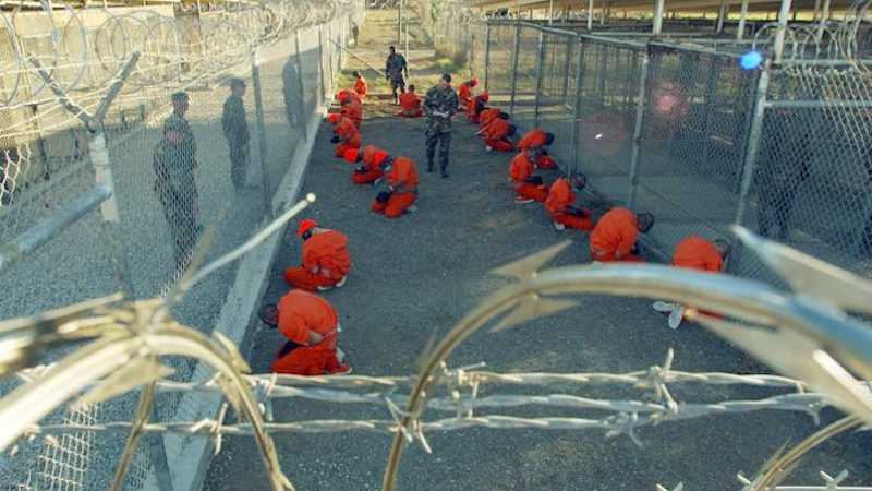 Detainees in orange jumpsuits sit in a holding area under the watchful eyes of Military Police at Camp X-Ray at Naval Base Guantanamo Bay, Cuba, during in-processing to the temporary detention facility on Jan. 11, 2002. DoD photo by Petty Officer 1st class Shane T. McCoy, U.S. Navy.